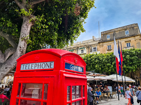 Valetta, Malta, May 12th 2023 - Red British-style telephone booth on the busy Republic Square piazza in Valetta, capital of Malta. People are sightseeing and enjoying in sidewalk cafes.