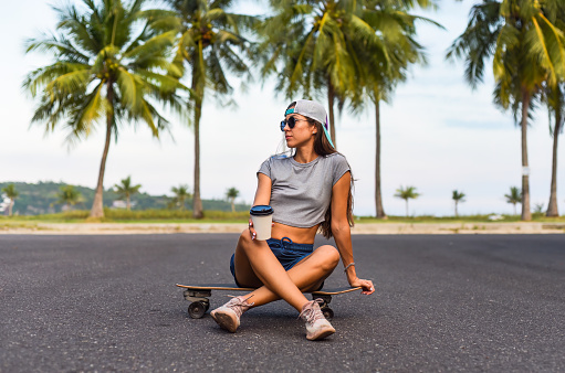 Young beautiful woman in a cap sitting on a skateboard and drinking coffee