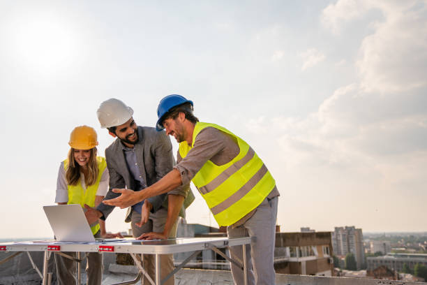 Three construction professionals in safety gear smile while reviewing plans on a laptop at a rooftop site.