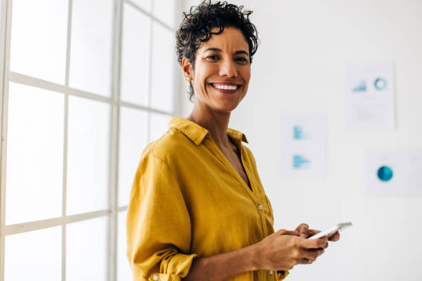 Happy female professional holding a mobile phone in her hand Happy female professional holding a mobile phone in her hand. Business woman browses business contacts on her phone as she stands next to a window in an office. small office interior stock pictures, royalty-free photos & images