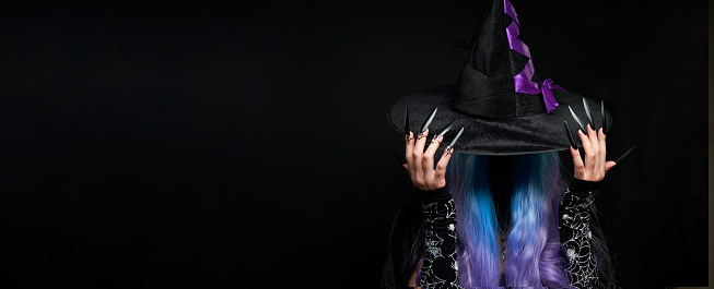 Halloween witch woman in black dress, cloak, black hat with violet ribbon, blue and violet hair and long black nails. Halloween celebration concept