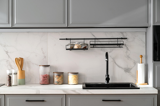 Front view of marble countertop with black sink near light ceramic wall and gray furniture in nordic style kitchen. Various jars with food and cooking utensils on stone surface.