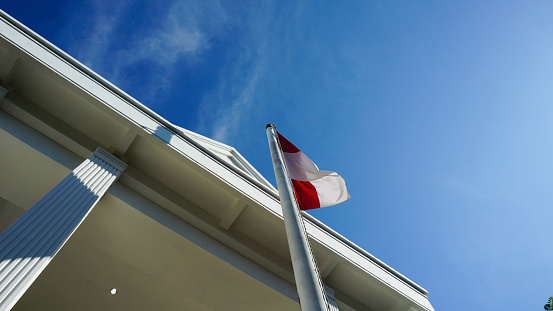 Indonesia flag fluttering on a pole in front of the building. Indonesian state flag, red and white. Blue and clear sky.
