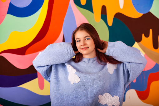 Happy teen lady model posing at colored wall, smiling looking at camera. Portrait of lovely teenage cover girl in blue fluffy sweater, positive emotion. Teenager emotional concept. Copy ad text space