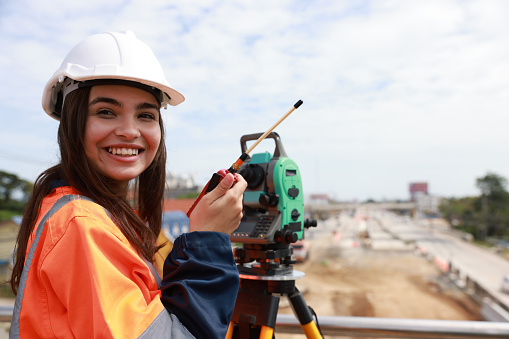 Site female engineer operating her instrument during roadworks looking at camera. Builder using total positioning station and looking at camera