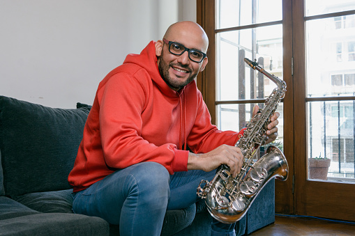 portrait of latino musician man dressed in red, with glasses and beard, at home sitting in the living room looking at the camera smiling happily with his saxophone with a large window in the background.
