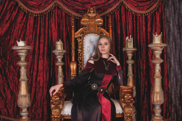 istock Medieval queen in historical image posing sitting on golden throne in castle room, looking at camera. Lovely lady in an old style on antique throne fortress. Concept of theatrical. Copy ad text space 1550860104