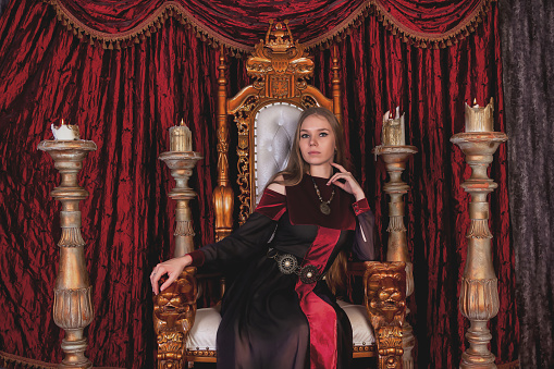 Medieval queen in historical image posing sitting on golden throne in castle room, looking at camera. Lovely lady in an old style on antique throne fortress. Concept of theatrical. Copy ad text space