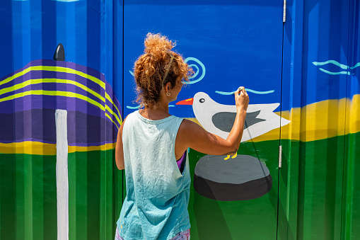 Mid adult Iranian woman artist painting  mural on shipping container, being converted in to public recreational facility.  She is dressed in casual work clothes. Exterior of public park and marina on the shore of canal in small town in Ontario, Canada.