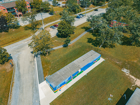 Mid adult Iranian woman artist painting  mural on shipping container, being converted in to public recreational facility.  She is dressed in casual work clothes. Exterior of public park and marina on the shore of canal in small town in Ontario, Canada. View from the drone.