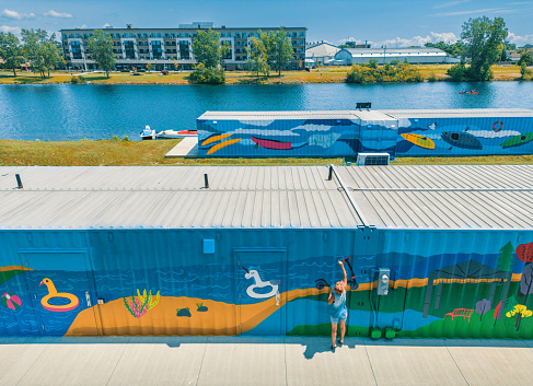 Mid adult Iranian woman artist painting  mural on shipping container, being converted in to public recreational facility.  She is dressed in casual work clothes. Exterior of public park and marina on the shore of canal in small town in Ontario, Canada. View from the drone.