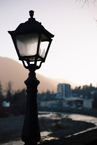 Capture the mesmerizing sunset view of Borjomi, Georgia, as the sun casts a warm glow on the city and mountains. In the foreground, an old street lamp stands as a nostalgic symbol of the past.