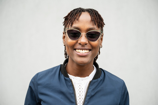 Close view of late 20s woman wearing navy zip-up jacket over white top, sunglasses, short locs hairstyle, and smiling at camera against white background.