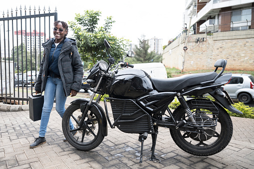 Candid full length portrait of smiling woman in casual clothing carrying mobile LFP battery from electric bike to off-camera automated swap cabinet, Nairobi.