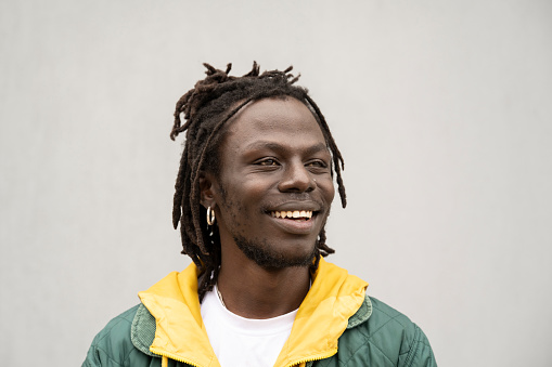 Close view of early 30s man with facial hair, locs hairstyle, wearing yellow and green jacket over t-shirt, looking away and smiling.