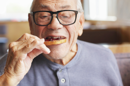 A senior man enjoys a bite of his toasted bread spread with sweet jelly jam preserves during breakfast at a restaurant. Selective focus on the toast and jelly.