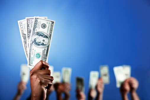One hand holds up a bundle of dollars which are spotlit by a heavenly beam of light, while many more hands do the same in the background against a deep blue background with plenty of copy space. 