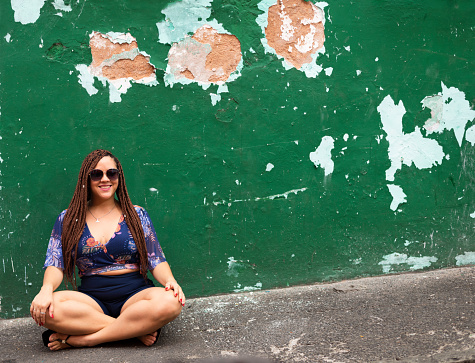 A woman sitting on the sidewalk wearing sunglasses and looking at the camera against an old green colored wall. Tourist on trip. Pelourinho, Salvador, Brazil.