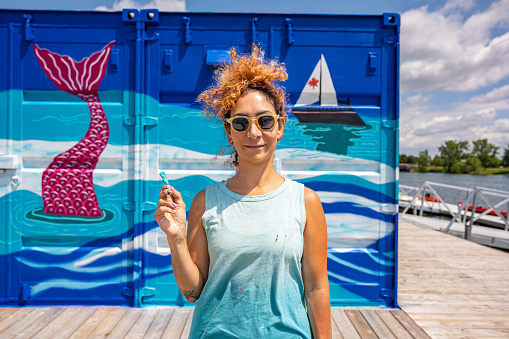 Mid adult Iranian woman artist painting  mural on shipping container, being converted in to public recreational facility.  She is dressed in casual work clothes. Exterior of public park and marina on the shore of canal in small town in Ontario, Canada.
