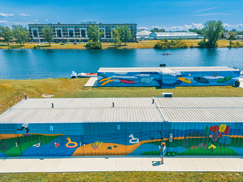 Areal view of Mid adult Iranian woman artist painting  mural on shipping container, being converted in to public recreational facility.  She is dressed in casual work clothes. Exterior of public park and marina on the shore of canal in small town in Ontario, Canada. View form the drone.