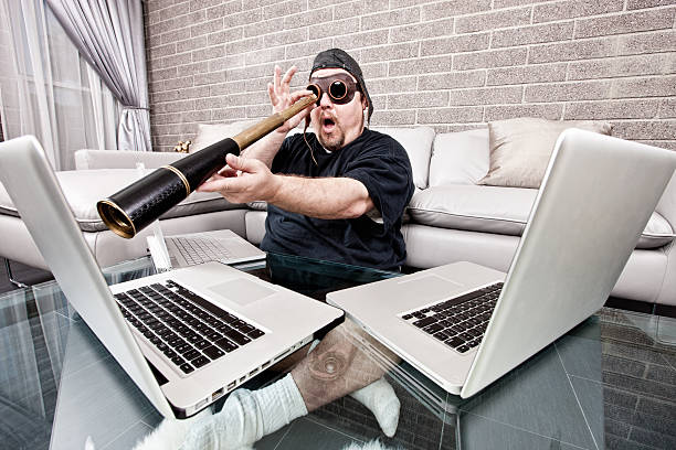 Techno Pirate with Spyglass Funny photograph of a man with three laptops sitting on the floor of his living room looking at the monitor through a hand-held telescope with a look of surprise on his face. Internet security, computer surveillance, remote network support, and identity theft concepts. computer hacker spy computer crime laptop stock pictures, royalty-free photos & images