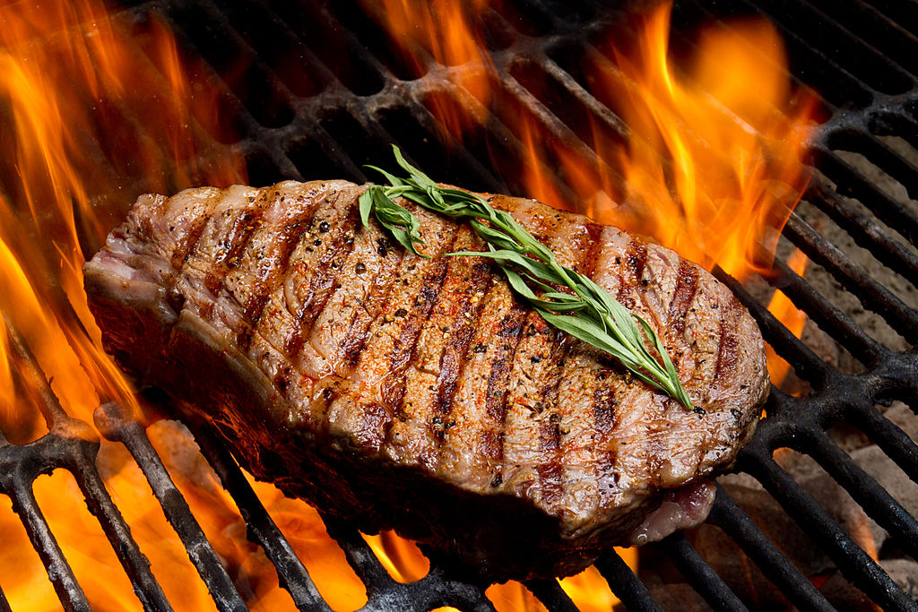Juicy ribeye steak on the grill kissed by fire on an old fashioned charcoal barbecue.  The steak is garnished with a sprig of rosemary and the grill marks are perfect