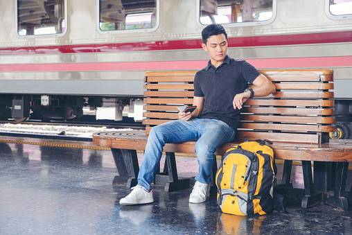 Cheap Travel and Lifestyle concept.Holiday Time,Young Solo Traveler man wearing sneaker and sitting at train station.Asian Backpacker waiting train alone and plan trip in summer time with smart phone