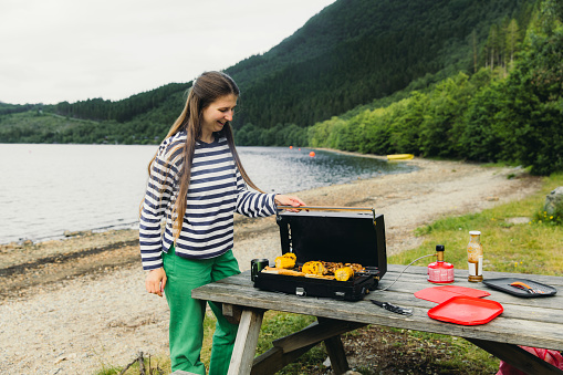 Side View of smiling female with long hair in striped shirt grilling chicken and corn on a grill stove by the lakeshore with view of beautiful green mountains
