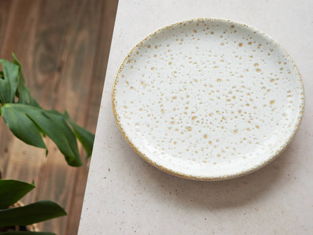 istock Empty white ceramic plate with gold dots on table, wooden floor and green plant leaf. Background with copy space. 1551051348