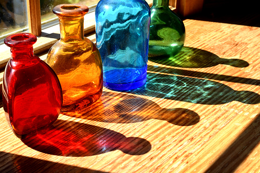 Four (4) different colored bottles on windowsill casting colored shadows from the sun.