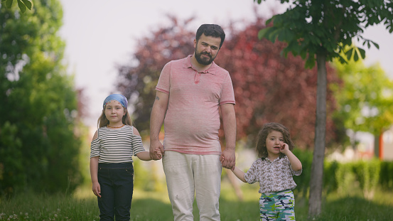 A portrait of a father and his daughter and son holding hands in a public park.