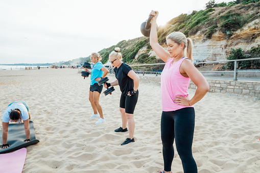 Active women of various ages doing fitness workouts in class exercise with coach on the beach. Ladies working with dumbbells and doing plank. Sport for health and wellbeing. Active lifestyle.