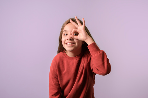 Girl shows peeping keyhole gesture symbol. Lilac background, studio portrait