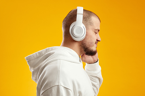 Side view of a cheerful young caucasian man listening to music with wireless headphones isolated over yellow background, wearing white hoodie