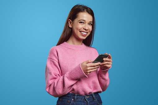 Cheerful hispanic female in pink sweater looking at camera with smile while using mobile app on smartphone, isolated against blue background