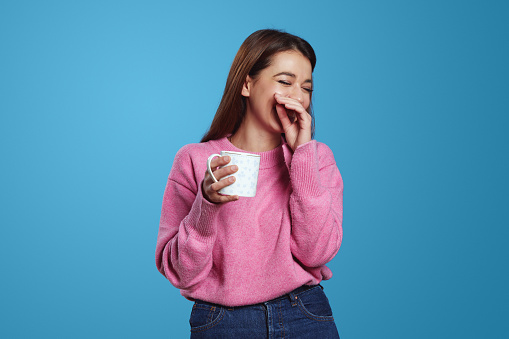 Portrait of brunette Hispanic girl with shy expression smiles pleasantly with cup of coffee wears pink jumper, isolated over blue background. People, emotions concept