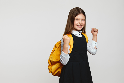 Excited schoolgirl in uniform and yellow backpack standing with fists up and celebrating achievement on white background. Back to school concept