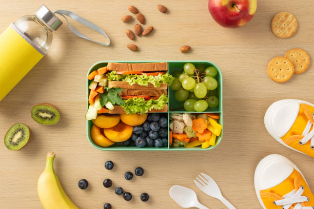 istock On wooden desk, plastic lunchbox is filled with nutritious elements such as kiwi, apple, banana, peach, grapes, vegetables, sandwich, blueberries, almonds, cookies, water bottle, cutlery, and sneakers 1551173260