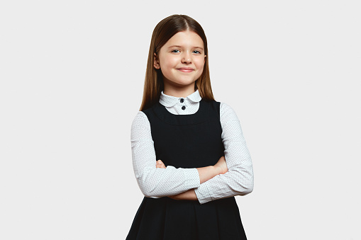 Smart focused girl in dress school uniform looking at camera while standing alone, with arms crossed against white background in studio