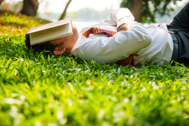 아시아 사업가는 잔디밭에 누워 팔짱을 끼고 낮잠을 자고 책으로 공원에서 점심 시간에 햇빛으로부터 눈을 보호하기 위해 얼굴을 가리고 있습니다. - resting relaxation book break 뉴스 사진 이미지