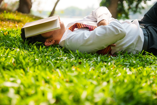 Asian businessman lying down on grass area and napping with arms crossed and book covering his face to protect eyes from sunlight during his lunch break at a park.