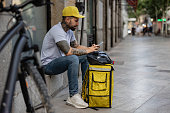 latino delivery man sitting with his cap, his backpack and bicycle in the foreground taking a break while looking at his mobile phone