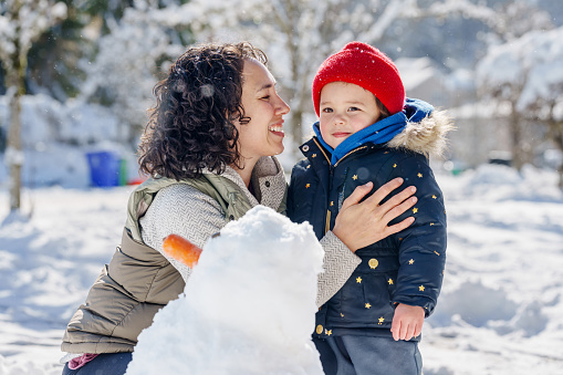 Multiracial mother of Hawaiian and Chinese descent smiles, looking at her preschool age daughter as they build a snowman together on a cold, winter day.