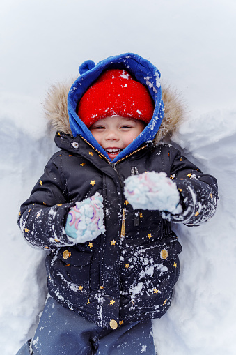 Multiracial preschool age girl of Chinese and Hawaiian descent smiles and laughs up toward the camera while laying on her back in the snow and making snow angel during a cold, winter day at home.
