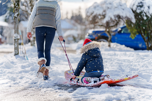 Preschool age girl sits on a sled while her mother pulls her along the sidewalk through their neighborhood on a snowy, winter day.