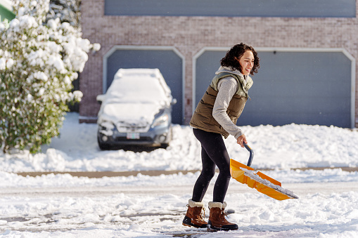 Young woman of Pacific Islander descent wearing warm winter clothes shovels snow off of her driveway during a cold, snowy day at home.