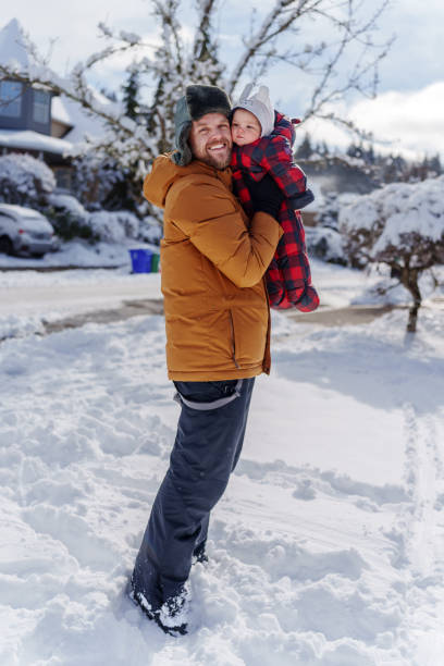 Young, white father holds his 6 month old son, smiling as they pose for a picture standing in the snow, bundled up in warm, winter clothing.