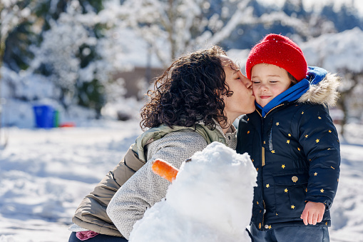 Young Eurasian mother kisses her preschool age daughter on the cheek as they build a snowman together on a cold, winter day.
