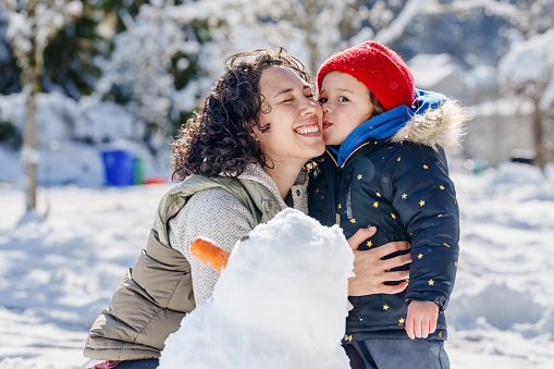 Preschool age Eurasian girl kisses her mother on the cheek as they build a snowman together on a cold, winter day.