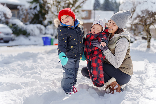 Multiracial mother of Pacific Islander descent and her preschool age daughter, and 6 month old son giggle and smile as they play together outside during a snowy, cold, winter day at home.